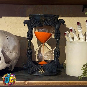 May include: Black hourglass with raven wings and a pentagram on the base. The hourglass is filled with orange sand.