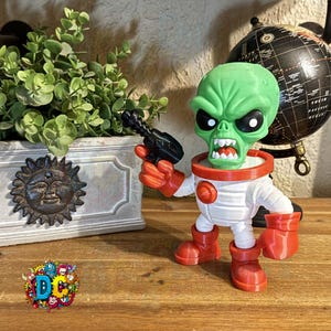 Puede incluir: Figura de alienígena verde con traje espacial blanco, guantes y botas rojas, sosteniendo una pistola de rayos negra. El alienígena tiene una expresión enfadada. Un sol decorativo y un globo terráqueo están en el fondo.