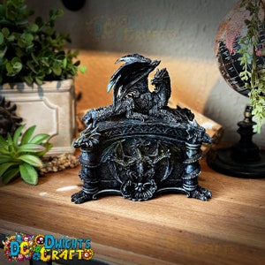 May include: A black dragon-shaped trinket box with a detailed design. The box is made of resin and features a dragon with wings spread out on top. The box is closed and has a decorative design on the front.