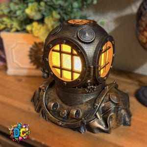 Peut inclure: Lampe en forme de casque de plongée de couleur bronze, avec une lumière jaune chaude émanant de la plaque frontale. La lampe présente des détails complexes, notamment des rivets, des hublots circulaires et une finition texturée, créant une esthétique antique.