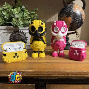 May include: A selection of yellow and pink protective cases for wireless earbuds, each designed as a hazmat suit figure. The cases are presented on a wooden surface, with a globe and decorative items in the background.