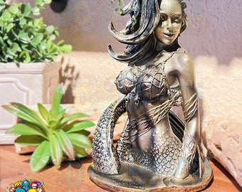 May include: A gold-toned mermaid statue with a detailed, scaly tail and flowing hair. The statue is posed with her head turned to the side and her hand resting on her tail. The statue is sitting on a base that resembles a wave.