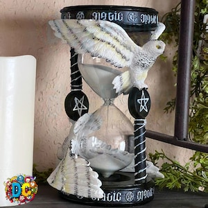 Puede incluir: Un reloj de arena negro con un búho blanco posado en la parte superior. El búho tiene las alas extendidas y un pentagrama a cada lado del reloj de arena. El reloj de arena está decorado con la palabra "magic" en la parte superior e inferior.