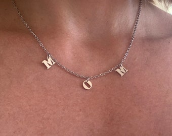 Puede incluir: Un collar de cadena de plata con tres dijes de plata. Los dijes son las letras "M", "O" y "M".