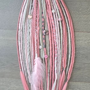 Puede incluir: Un conjunto de rastas sintéticas de color rosa, blanco y gris con varios adornos, incluyendo cuentas, botones y encaje.
