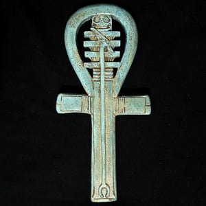 May include: A light blue and gold ankh symbol featuring a central djed pillar and a cobra figure. The object has a weathered, textured finish with a prominent loop at the top, horizontal crossbar, and a long vertical base stem.