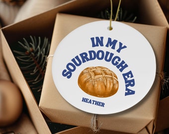 Könnte beinhalten: Ein weißer, runder Weihnachtsschmuck mit dem Text "IN MY SOURDOUGH ERA" in Blau und einer Grafik eines Brotlaibs. Der Name "HEATHER" steht unter der Brotgrafik. Der Schmuck befindet sich in einer braunen Geschenkbox mit einem goldenen Faden.