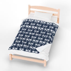 May include: A navy blue blanket featuring a white ping pong paddle and laurel wreath design. The blanket is on a small wooden bed with white sheets and a pillow. The bed frame is made of light-coloured wood.