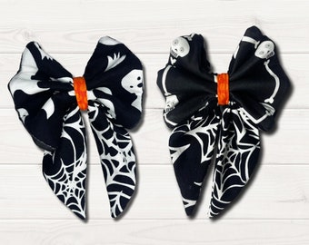 May include: Two black and white fabric hair bows with a spiderweb and skeleton design. The bows are tied with an orange elastic band.
