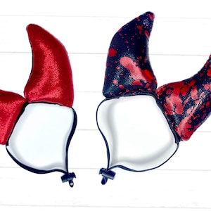 May include: Two pairs of horn-shaped head accessories with adjustable black elastic straps. One pair is solid metallic red, while the other pair features a black base with a red splatter pattern. Both sets have white interior linings and black toggles.