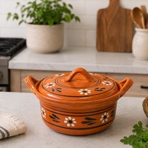 May include: An orange ceramic cooking pot with a lid, adorned with white floral and black leaf designs. The pot has two side handles and a top handle. It is sitting on a countertop in a kitchen setting.