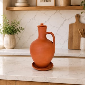 May include: A terracotta-colored ceramic pitcher with a lid and handle, resting on a matching saucer. The pitcher has a rounded body and a narrow neck. The kitchen setting includes a wooden cutting board and a vase with utensils.