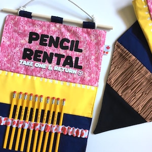 Op de afbeelding: Een stoffen wandhanger met een roze bloemmotief aan de bovenkant en de zwarte tekst PENCIL RENTAL TAKE ONE & RETURN. Daaronder een gele strook met de cijfers 1 tot en met 18 en een donkerblauw gedeelte met een rij gele potloden in vakjes.