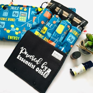 Puede incluir: Una bolsa negra con cremallera con el texto "Powered by Essential Oils" en blanco. La bolsa tiene un forro de tela azul y naranja con un tema científico, con vasos de precipitados, moléculas y las palabras "Ionic Bond", "Joule" y "Energy". La bolsa está abierta y muestra cuatro pequeños frascos de aceites esenciales en el interior.