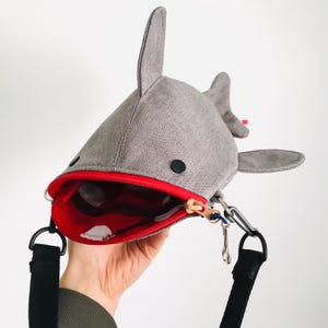 May include: Gray shark-shaped crossbody bag with a red interior and zipper. The bag has a black strap and a small, red accent near the fin. The shark's mouth is open, revealing a red interior with white polka dots.