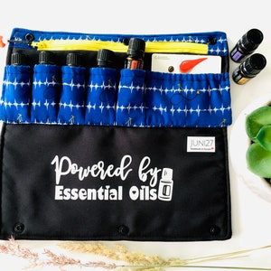 Puede incluir: Una bolsa de tela azul y negra con múltiples bolsillos para botellas de aceites esenciales. La bolsa tiene un cierre de cremallera y una etiqueta blanca que dice "Powered by Essential Oils".