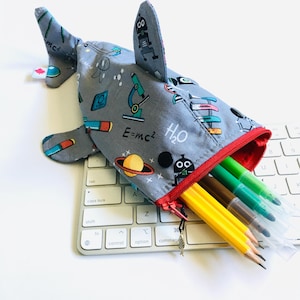 Peut inclure: Une trousse à crayons en tissu gris en forme de requin, avec un motif sur le thème des sciences incluant des microscopes, des planètes et le texte E=mc2 et H2O. Elle possède une fermeture éclair rouge avec une breloque poisson et des crayons.