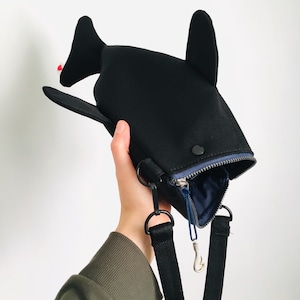 May include: A black whale-shaped crossbody bag with a zipper closure and a blue interior. The bag has a black adjustable strap and a silver-colored clasp. The whale has fins and a small red detail.
