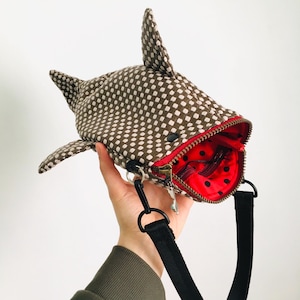 May include: A novelty crossbody bag shaped like a whale shark. The bag is brown with white polka dots, featuring fins and a tail. The interior is red with black polka dots, and it has a black strap and zipper.
