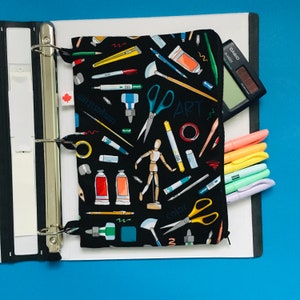 Könnte beinhalten: Eine schwarze Reißverschlusstasche mit einem bunten Kunst-Motiv mit Pinseln, Scheren und dem Wort "ART". Die Tasche ist offen und befindet sich in einem Ordner mit einem Taschenrechner und Buntstiften.