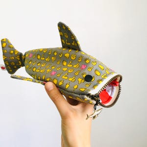 Peut inclure: Une trousse en tissu en forme de requin de couleur grise avec un motif répétitif d'emojis de gestes de la main jaunes. La trousse présente une bouche grande ouverte avec une fermeture éclair en métal, un œil circulaire noir et une breloque.