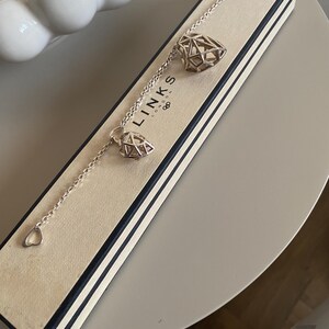 Op de afbeelding: Een zilveren armband met geometrische diamantvormige bedels en hartvormige details, gepresenteerd in een merkdoos. De armband heeft een fijne ketting en wordt gepresenteerd op een neutrale ondergrond. De doos heeft zwart-witte strepen.