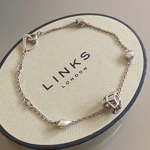 Op de afbeelding: Zilveren armband met hart- en geometrische kubusbedels. De armband ligt op een beige doos met de merknaam "LINKS LONDON". De armband heeft een fijne ketting en verschillende hartvormige en geometrische bedels.
