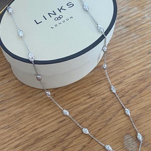 Puede incluir: Un collar plateado con cuentas redondas y ovaladas texturizadas alternas. El collar se muestra sobre una caja crema con el logotipo "LINKS LONDON". El collar está sobre una superficie de madera.