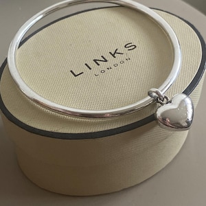 Puede incluir: Una pulsera de plata con un dije de corazón descansa sobre una caja beige. La pulsera es circular y tiene un acabado pulido. El dije de corazón tiene forma redondeada. La caja tiene la marca "LINKS LONDON" impresa.