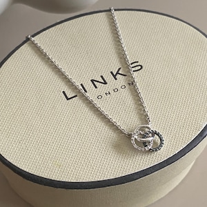 May include: A silver chain necklace featuring a pendant with two interlocking circular rings, one set with clear stones and the other with dark stones, resting on a cream-coloured oval box labelled with the text LINKS LONDON in black lettering.