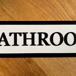 May include: A rectangular bathroom sign with a white background and black border. The word "BATHROOM" is prominently displayed in bold, black letters. The sign has a decorative, curved edge.