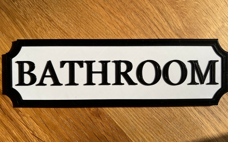 May include: A rectangular bathroom sign with a white background and black border. The word "BATHROOM" is prominently displayed in bold, black letters. The sign has a decorative, curved edge.