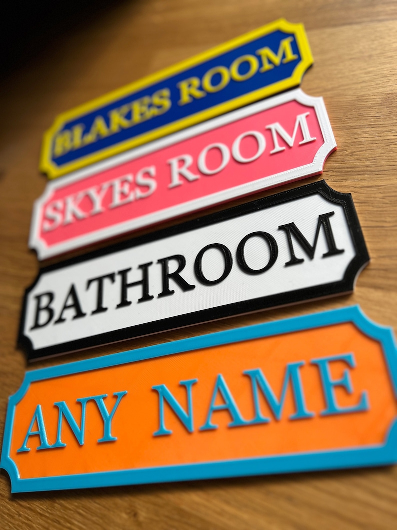 May include: A stack of colorful room signs with raised lettering. The signs include "BLAKES ROOM" in blue and yellow, "SKYES ROOM" in pink, "BATHROOM" in black and white, and "ANY NAME" in orange and blue. The signs are on a wooden surface.