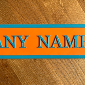May include: A rectangular sign with the words "ANY NAME" in blue, raised letters. The sign has an orange center and a blue border. The sign is on a wooden surface.