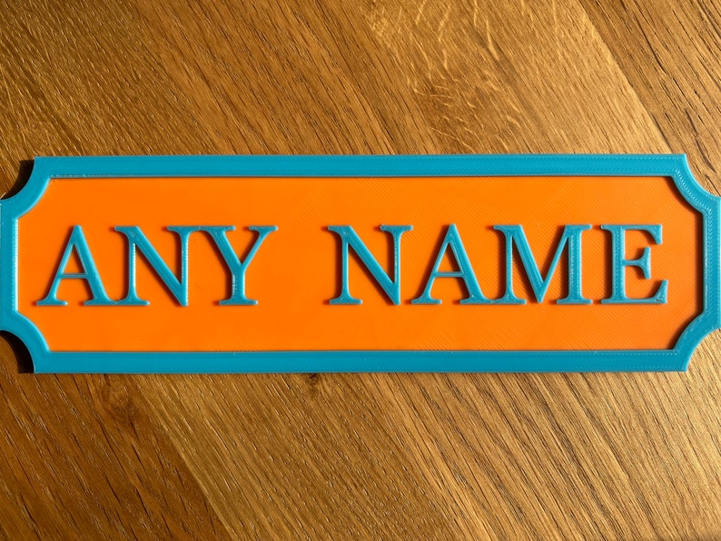 May include: A rectangular sign with the words "ANY NAME" in blue, raised letters. The sign has an orange center and a blue border. The sign is on a wooden surface.