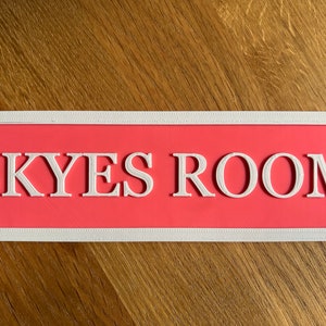 May include: A rectangular sign with the text "SKYES ROOM" in white, set against a coral background. The sign has a white decorative border. The sign is likely for a bedroom or living space.