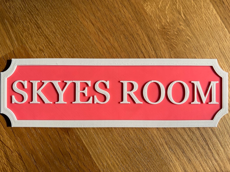 May include: A rectangular sign with the text "SKYES ROOM" in white, set against a coral background. The sign has a white decorative border. The sign is likely for a bedroom or living space.