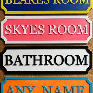 May include: Four colorful room signs with raised lettering. The top sign is blue with yellow trim and reads "BLAKES ROOM." Below it is a pink sign with white trim that reads "SKYES ROOM." The third sign is white with black trim and reads "BATHROOM." The bottom sign is orange with blue trim and reads "ANY NAME."