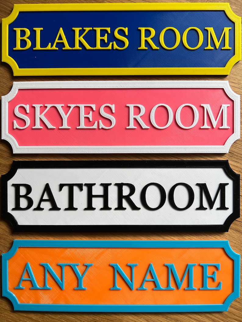 May include: Four colorful room signs with raised lettering. The top sign is blue with yellow trim and reads "BLAKES ROOM." Below it is a pink sign with white trim that reads "SKYES ROOM." The third sign is white with black trim and reads "BATHROOM." The bottom sign is orange with blue trim and reads "ANY NAME."