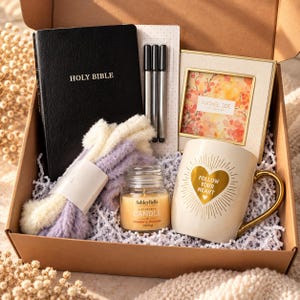 May include: A gift box containing a black Holy Bible, three black pens, a Rachel Zoe picture frame with floral art, a pair of fuzzy lavender and cream socks, a small Ashley Belle strawberry shortcake scented candle, and a cream mug with gold text.