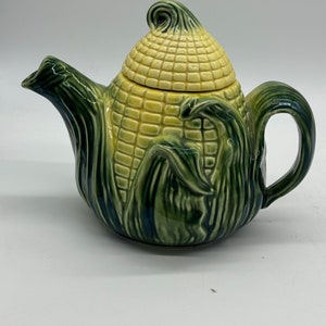 Peut inclure: Une théière en céramique en forme d'épi de maïs. La théière est principalement jaune et verte, avec une surface texturée imitant les grains et les feuilles de maïs. La poignée et le bec sont verts, et le couvercle est d'un jaune plus clair.