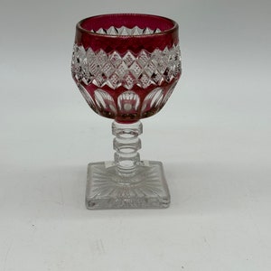 May include: A clear crystal wine glass with a square base and a stem featuring stacked rings. The rim of the glass is a deep red colour, and the bowl has a diamond-cut pattern.