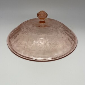May include: A pink glass lid with a floral pattern and a small, faceted knob. The lid is round and appears to be part of a serving dish or bowl. The glass has a delicate, translucent quality.