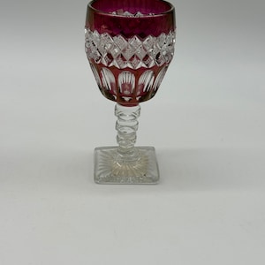 May include: A clear glass wine goblet with a square base and a decorative stem. The top of the goblet is a deep burgundy colour, with a diamond-cut pattern around the rim. The goblet is designed for serving wine.