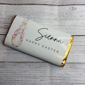 May include: A chocolate bar with a white wrapper featuring a bunny illustration in a pink jacket. The name "Sienna" and the words "Happy Easter" are printed on the wrapper, a seasonal Easter treat.