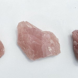 May include: Three pink rose quartz crystals of varying sizes and shapes. The crystals are rough and have a translucent appearance.