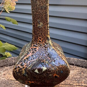 May include: A unique, handcrafted ceramic vase with a textured, mottled surface in shades of brown, black, and orange. The vase has a bulbous base, a long neck, and a flared top. The design includes a stylized eye.