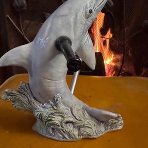 May include: A ceramic dolphin figurine with a blue and gray color scheme. The dolphin is holding a black metal rod in its mouth. The figurine is sitting on a green and brown base that resembles seaweed.
