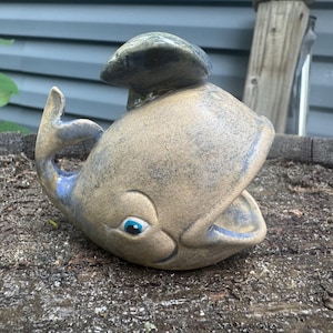 May include: A ceramic whale figurine with a whimsical design. The whale is primarily tan with blue accents on the fin and mouth. It has a small, round, blue eye and a spout on top. The figurine is sitting on a weathered wooden surface.