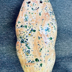 May include: A smooth, oval-shaped ceramic figurine with a glossy finish, featuring a speckled pattern of tan, dark green, and blue spots. The object has two small dark circular indentations for eyes and a curved line for a mouth, creating a face.